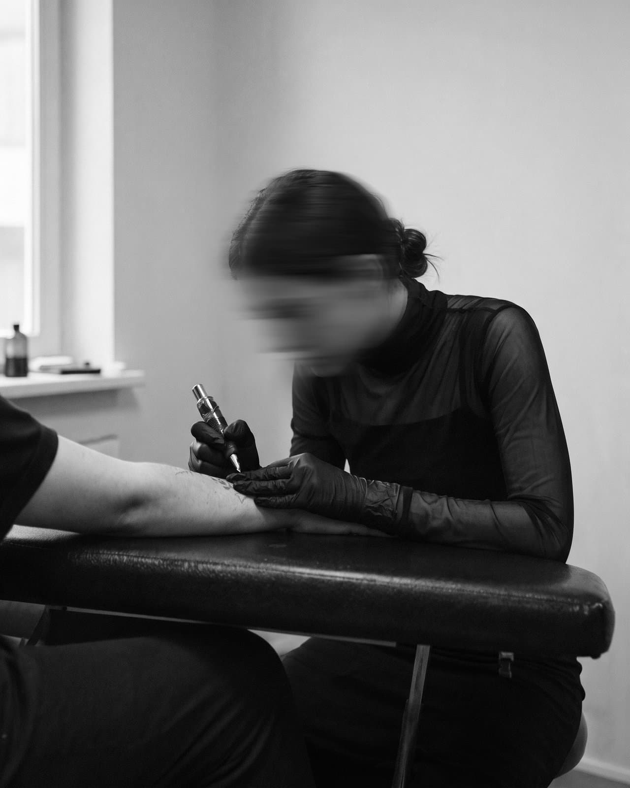 A tattoo artist in black gloves works on a client's forearm, her face blurred in motion